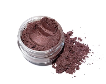May include: A small jar of loose brown eyeshadow with a shimmery finish. The eyeshadow is spilled out around the jar, showing the texture and color.