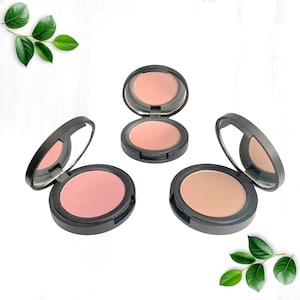 May include: Three open compacts of blush makeup in shades of pink and peach. Each compact has a black case with a built-in mirror. The blush colors range from a light peach to a deeper rose. Green leaves are in the background.