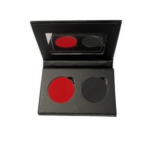 May include: A black makeup palette with a built-in mirror. The palette contains two circular pans, one filled with bright red product and the other with a matte black product. The palette is open, revealing the contents.