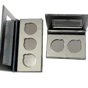 May include: Two open, black eyeshadow palettes with silver inserts. One palette has three circular wells, the other has two. Each palette has a clear mirror in the lid. The wells are empty, ready to be filled with eyeshadow.