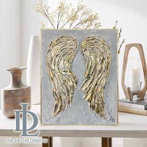 May include: A gray canvas painting with two gold textured angel wings. The wings are raised and spread out. The painting is on a white surface.