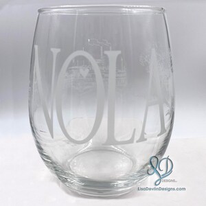 May include: A clear glass wine glass with the word "NOLA" etched in white. The glass also features a small etched image of a steamboat with a heart.