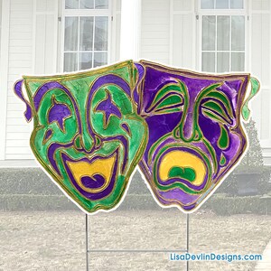 May include: Two colorful Mardi Gras masks, one green and gold with a smiling face and the other purple and gold with a crying face. The masks are attached to a metal stake for outdoor display.