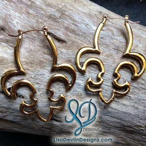 May include: A pair of gold-toned hoop earrings with a decorative fleur-de-lis design. The earrings are made of metal and have a shiny finish.
