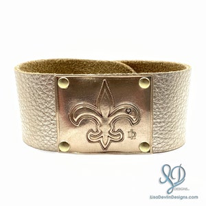 May include: A gold leather bracelet with a gold metal fleur-de-lis charm. The charm has a small heart in the center and the letters "ID" below it.