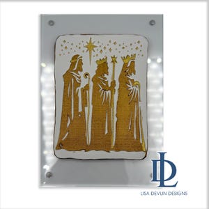 May include: A gold-colored depiction of the Three Wise Men, with a star and celestial accents, is presented on a white background. The artwork is framed in a clear acrylic sheet, secured with silver-colored screws. The artist's name, Lisa Devlin Designs, is printed at the bottom.