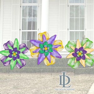 May include: Three colorful, handcrafted flower garden stakes. Each flower features layered petals in shades of green, purple, and yellow, with a contrasting center. The stakes are designed for outdoor use, adding a decorative touch to any garden or yard.