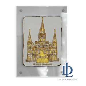 May include: A framed, rectangular artwork featuring a gold-colored relief of St. Louis Cathedral on a white background. The artwork is encased in a clear acrylic frame. The text "ST. LOUIS CATHEDRAL" is visible at the bottom.