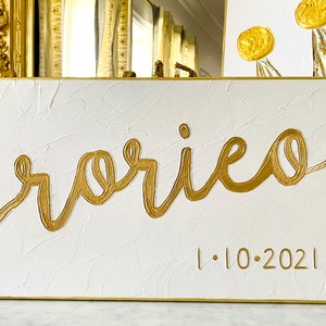 May include: A rectangular white canvas with textured surface, featuring the gold cursive word "rorieo" and the date "1.10.2021". The artwork has a gold border and is likely a personalized piece.