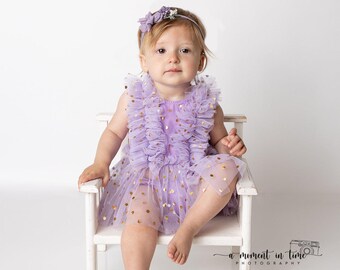 May include: A lavender tulle dress featuring vertical ruffles on the bodice and a skirt adorned with metallic gold polka dots. The garment is paired with a matching lavender floral headband, worn by a child sitting on a small white wooden chair.