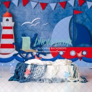 May include: A white box with blue and white netting, a red starfish, and a blue starfish. The box is on a white surface in front of a blue and red nautical themed backdrop with a lighthouse, a sailboat, and a wheel.