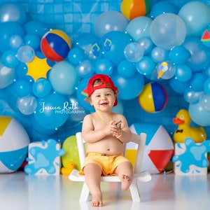 May include: A young child wearing a red baseball cap and yellow swim trunks sits on a white chair in front of a blue and white balloon arch. The balloons are decorated with beach-themed icons like flip flops, ice cream cones, and watermelons. The child is smiling and holding a small object in their hands. The background is a blue tiled wall.