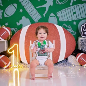 May include: A baby boy wearing a white onesie with a green football design and the name "Cristian" on it. He is sitting on a wooden chair in front of a green and brown football-themed backdrop. A neon yellow number one is in the foreground.