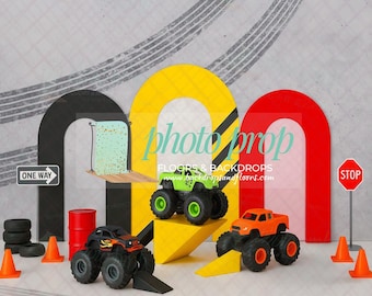 May include: A photography backdrop featuring toy monster trucks, traffic cones, and a stop sign. The backdrop includes a gray background with tire tracks, and colorful arches in yellow, black, and red. The text "photo prop" is visible.