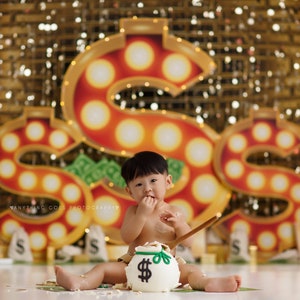 May include: A baby sitting in front of a money themed backdrop with a large dollar sign made of lights. The baby is holding a cake shaped like a money bag with a dollar sign on it.