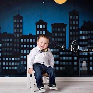 May include: A young boy wearing a white shirt and blue jeans sits on a white chair in front of a city skyline backdrop. The backdrop is a dark blue with a yellow moon and stars. The boy is wearing a bow tie with a superhero design.