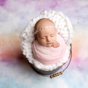 以下が含まれることがあります： 生まれたばかりの女の子が、白い編み物のバケツの中で、ピンクの包みに包まれてぐっすり眠っています。赤ちゃんは、金の星がついたヘッドバンドをしています。背景は、カラフルな水彩画風の背景に、金のキラキラした星が散りばめられています。
