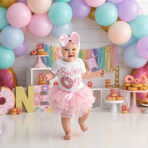 May include: A baby girl wearing a pink tutu and a white shirt with the text "Sweet One" and a donut graphic is standing in front of a donut-themed birthday party backdrop. The backdrop includes a rainbow banner, a donut-shaped inflatable, and a large gold "ONE" sign.