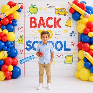 May include: A young boy stands in front of a backdrop with the words "Back to School." The backdrop is decorated with school-themed drawings and balloon arches in red, yellow, and blue. The boy holds an apple.