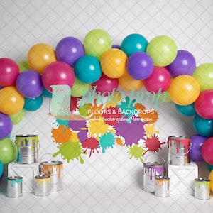May include: A colorful balloon arch made of pink, purple, yellow, green, and blue balloons. The arch is in front of a white backdrop with paint splatters and paint cans.