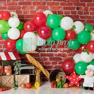 May include: A red brick backdrop with a balloon arch made of red, white, and green balloons. There is a small wooden market stand with red and white striped awning, a pasta maker, and a chef doll in front of the backdrop.
