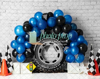 May include: A blue and black balloon arch with checkered flags and a tire-shaped inflatable. The backdrop is a white brick wall. The text "photo prop" is visible in the image.