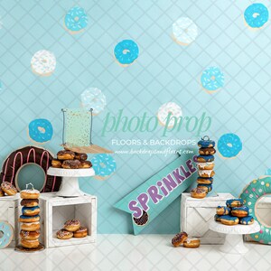 May include: A blue and white donut themed backdrop with white crates and cake stands holding stacks of doughnuts. A large blue and white donut ring is on the right side of the image. A sign reads "Sprinkle".