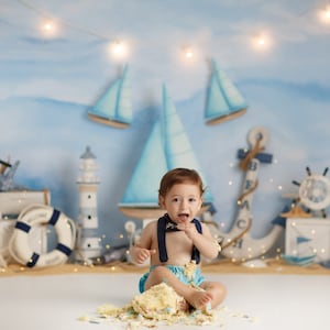 May include: A baby boy wearing a blue and white sailor outfit sits on a white surface with a messy cake in front of him. He is smiling and has cake on his face. The background is a blue and white nautical themed backdrop with sailboats, anchors, and lighthouses.