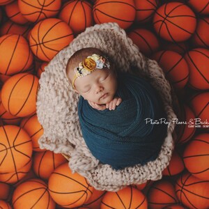 May include: A newborn baby girl is swaddled in a teal blanket and wearing a flower crown. The baby is lying in a basket surrounded by orange basketballs.