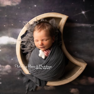 以下が含まれることがあります： 三日月型の木製小道具に寝ている新生児。グレーの包みに包まれ、白い星型の玩具を持っています。背景は濃い青と紫の星空です。