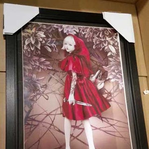 May include: A framed print of a young woman in a red hooded dress and holding a knife. The woman is standing in a woodland setting with a dark background.