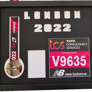 May include: A black framed display with a silver medal, a pink and black finisher's ribbon, a white rectangular space, and a pink and black race bib with the text "tcs TATA CONSULTANCY SERVICES tos #WeRunTogether V9635 new balance".