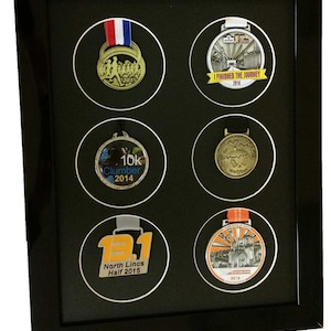 Puede incluir: Marco negro de exhibición de medallas con seis aberturas circulares, cada una con una medalla diferente. Las medallas son de plata, oro y bronce y presentan varios diseños y textos, incluyendo "I Finished the Journey 2016", "10k Clumber 2014" y "13.1 North Lincs Half 2015".