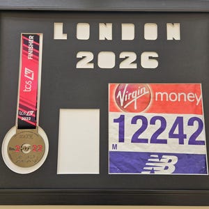 May include: A framed display featuring a London Marathon finisher's medal and race bib. The medal has a red and black ribbon and the year 2022. The race bib shows the number 12242 and the Virgin Money logo, with the year 2026 in the frame.