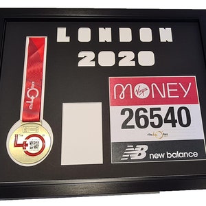 May include: A framed display of a London Marathon medal and race bib. The medal is gold with a red, white and blue ribbon. The bib is red and white with the text "Virgin Money" and the number "26540". The frame is black and has the text "LONDON 2020" in white.