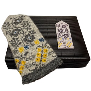 May include: A gray and white knitted mitten with a floral pattern in yellow and blue. The mitten is on top of a black box with a smaller gray and white knitted bookmark with a floral pattern.