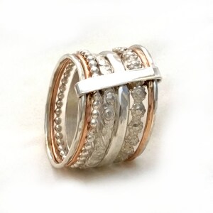 Puede incluir: Un anillo de plata y oro rosa con múltiples bandas, cada una con una textura diferente. Las bandas están apiladas e interconectadas, creando un diseño único e intrincado.