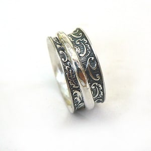 Puede incluir: Un anillo giratorio plateado con una banda central y dos bandas exteriores. Las bandas exteriores presentan un patrón oscuro y ornamentado de remolinos. El anillo está diseñado para girar, ofreciendo una experiencia táctil. El anillo se muestra sobre un fondo blanco.