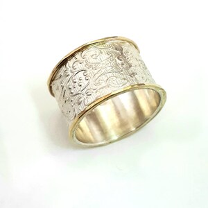May include: A wide silver ring with an ornate floral pattern. The ring has a gold band on the top and bottom edges. The ring is set against a white background.