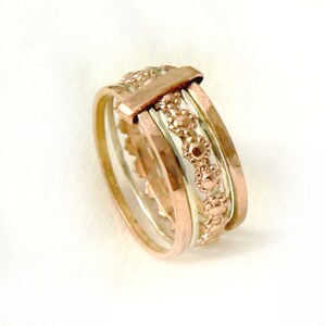 Puede incluir: Un anillo de oro con múltiples bandas, cada una con una textura diferente. La banda superior tiene una fila de pequeños círculos en relieve.