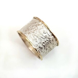 May include: A wide silver ring with an embossed floral pattern. The ring has gold-colored edges, creating a two-tone effect. The design features detailed, swirling floral motifs across the band.