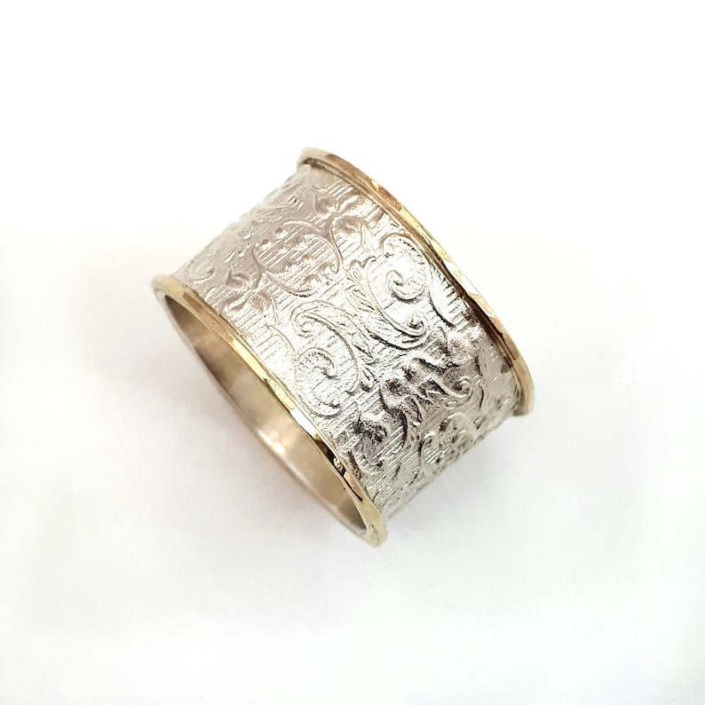 May include: A wide silver ring with an embossed floral pattern. The ring has gold-colored edges, creating a two-tone effect. The design features detailed, swirling floral motifs across the band.