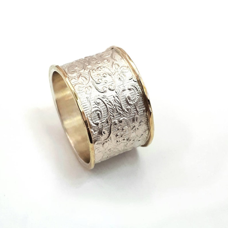 May include: A wide silver ring with an intricate floral pattern. The edges are trimmed with gold, creating a two-tone effect. The ring is set against a white background, highlighting its detailed design.