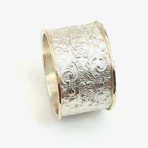 May include: A wide silver ring with an engraved floral pattern. The ring has gold-colored edges, creating a two-tone effect. The design is detailed and the ring appears to be made of metal. The ring is a piece of jewelry.