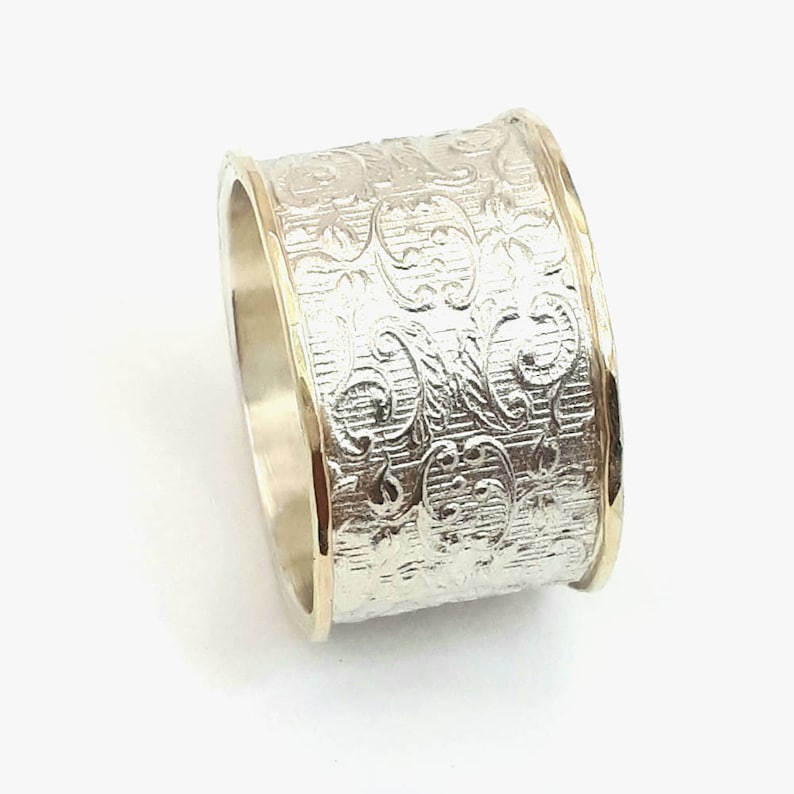 May include: A wide silver ring with an engraved floral pattern. The ring has gold-colored edges, creating a two-tone effect. The design is detailed and the ring appears to be made of metal. The ring is a piece of jewelry.