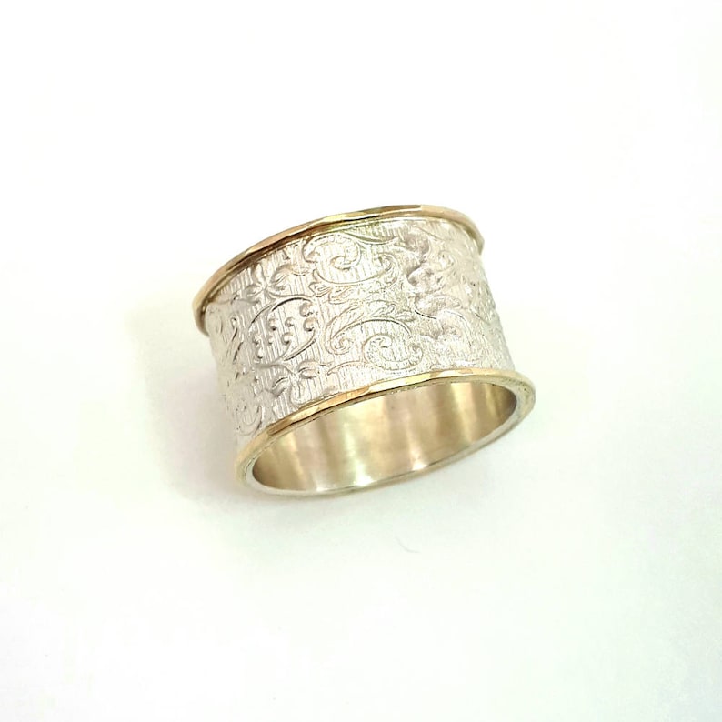 May include: A wide silver ring with an engraved floral pattern. The ring has gold-colored bands on the top and bottom edges. The ring is a statement piece of jewelry.