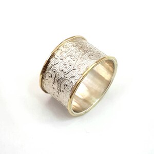 May include: A wide silver ring with an engraved floral pattern. The ring has gold-colored bands on the top and bottom edges. The ring is set against a white background.