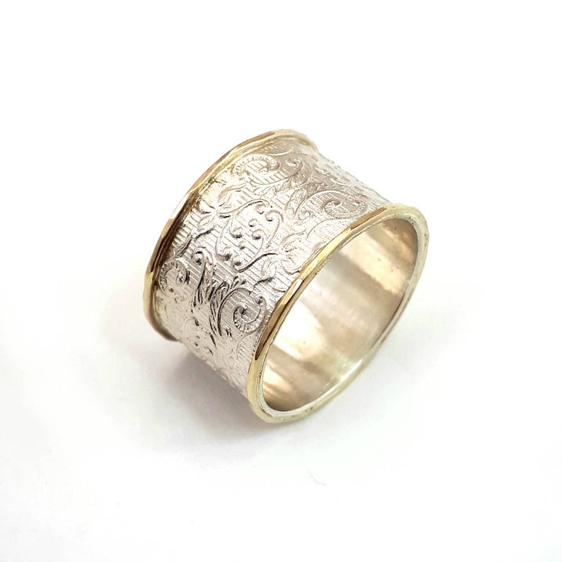 May include: A wide silver ring with an engraved floral pattern. The ring has gold-colored bands on the top and bottom edges. The ring is set against a white background.