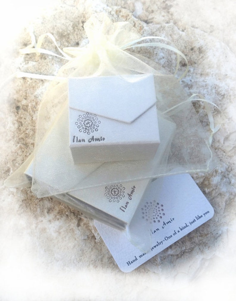 May include: A white jewelry box with a decorative design and the name "Ilan Amir" printed on it. The box is inside a sheer, off-white organza bag, with additional cards featuring the same design and text. The text reads "Hand made jewelry. One of a kind, just like you."