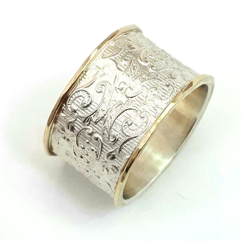 May include: A wide silver ring with an ornate, textured floral pattern. The edges are trimmed with gold, creating a two-tone effect. The ring's design features detailed scrollwork and a brushed metal finish.